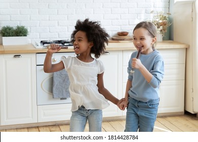 Funny Adorable Little Mixed Race Preschool Kids Enjoying Singing Songs In Kitchen Utensils As Microphones. Happy Overjoyed Small African Ethnicity Girl Having Fun With European Stepsister Or Friend.