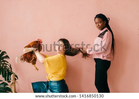Funny african american couple and caucasian girl with chicken in her hands standing all together on pink background in studio. Odd people concept.