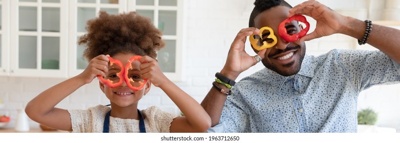 Funny African American dad and daughter girl making pepper slice glasses, having fun while cutting vegetables, cooking in kitchen together. Family eating at home, healthy food concept. Banner photo - Powered by Shutterstock