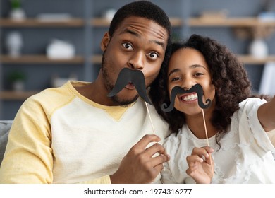 Funny african american father and daughter taking selfies together while staying home, using selfie accessories mustaches on sticks, looking at camera and smiling, closeup portrait - Powered by Shutterstock