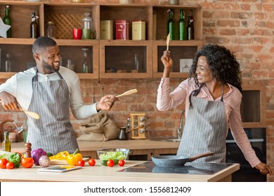 Funny African American Married Couple Fighting With Utensils Tools, Feeling Playful In Kitchen, Copy Space