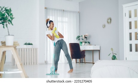 Funny African American teenager is cleaning house doing housework cleaning floor with mop and listening to music in wireless headphones, girl is dancing and singing. - Shutterstock ID 2231270405