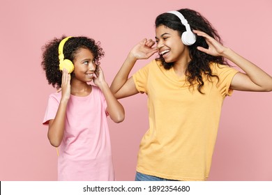 Funny African American Young Woman And Little Kid Girl Sisters In Casual T-shirts Listening Music With Headphones Looking At Each Other Isolated On Pink Background Studio Portrait. Family Day Concept