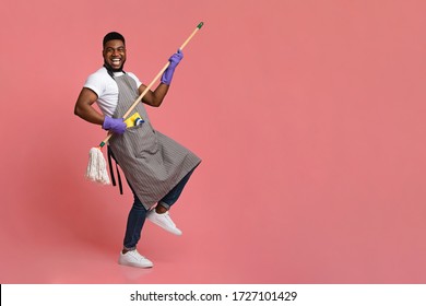 Funny African Man Using Mop Handle As Guitar, Having Fun During Cleaning. Posing Over Pink Background In Studio, Copy Space