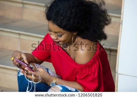 Funny african woman sitting on stairs listening to music with headphones and a smart phone outdoors.