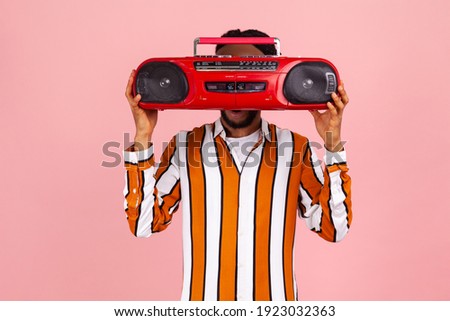 Funny afro-american man with beard and dreadlocks in bright striped shirt hiding his face behind red vintage audio record player, selling junk. Indoor studio shot isolated on pink background