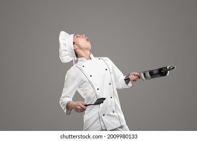 Funny amazed male chef in white uniform and hat holding frying pan and spatula and looking up, as juggling food during cooking process on gray background