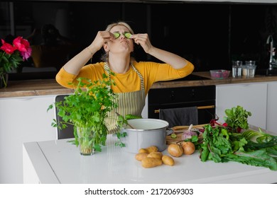 Funny, Amusing Blonde Woman In Apron Hold Cucumber Slices On Eyes As Mask In Kitchen. Fresh Vegetables For Soup Or Salad. Potato, Greenery, Beetroot Near Pan On Table. Cosmetology Treatment At Home