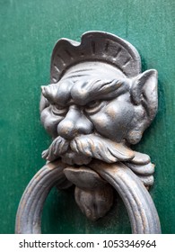 Funny, Amusing Or Humorous Close Up Photograph Of A Weathered Metal Antique Door Knocker Caricature Of A Man With A Mustache Big Ears And Nose On A Green Painted Wood Door In Pisa Italy.