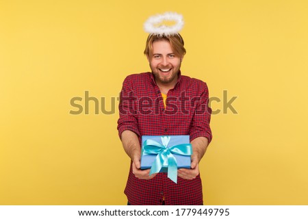 Funny angelic bearded guy with saint halo on head holding gift box, giving present and smiling kind friendly, concept of donation holiday charity. indoor studio shot isolated on yellow background