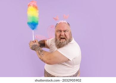 Funny Angry Bearded Plump Man In Fairy Suit With Wings Attacks With Fluffy Dust Brush On Purple Background In Studio