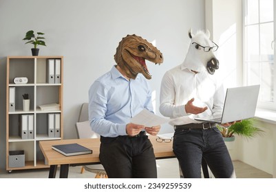 Funny animal people use computer at work. Team of 2 men in unusual masquerade dinosaur and horse masks standing by desk in office workplace, preparing creative business presentation on laptop device