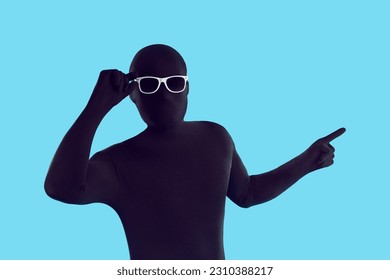 Funny anonymous faceless man disguised in monochromatic black skintight spandex bodysuit costume and sunglasses standing on blue background and pointing his finger away to show something on the side