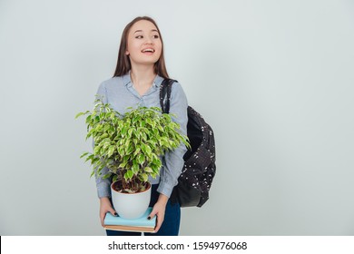 Funny Asain Student Girl Standing, Holding Two Books With A Flowerpot On The Top Of Them, Looking To The Side As If Wanting To See Someone. Backpack Slung Over Her Shoulder.