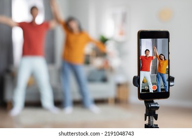 Funny Asian Husband And Wife Recording Video For Social Media Or Personal Family Vlog, Holding Hands And Dancing In Front Of Camera, Selective Focus On Modern Cell Phone On Tripod, Home Interior