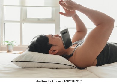 Funny Asian Man Let The Smartphone Fall Down To The Face While He Is Lying On Bed.