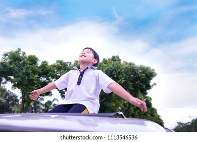 Funny Asian Young Boy Standing On The Sunroof Of The Car With Cloud,tree And Blue Sky Background.Smiling And Happiness Of Cute Kid In The Summer.Holiday,Vacation,Family,Travel Concept.