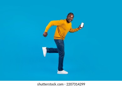 Funny Attractive Black Man In Casual Outfit Running Away With Modern Smartphone With White Blank Screen In His Hand, Blue Studio Background, Side View, Copy Space, Full Length Shot, Mockup