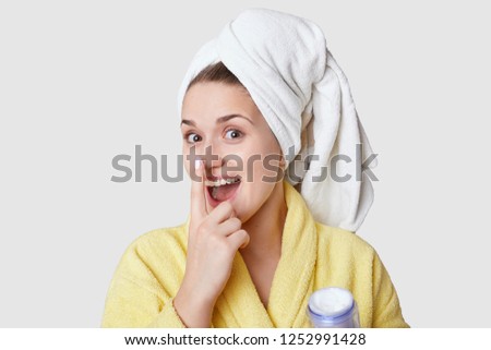 Funny attractive young woman keeps fore finger on nose, applies beauty cream, stands in bathroom, wears white towel and yellow robe, stands against white studio wall. Cosmetic and women concept