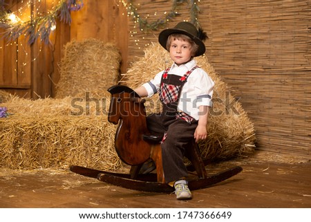 Funny baby boy sitting on the toy horse wearing retro costume in farm background with straw sheaves