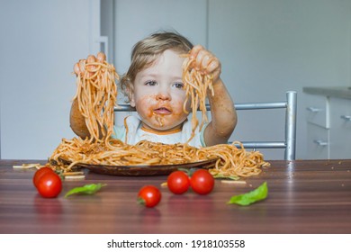 Funny Baby Child Getting Messy Eating Spaghetti With Tomato Sauce From A Large Plate, By Itself With His Hands, At Home