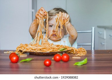 Funny baby child getting messy eating spaghetti with tomato sauce from a large plate, by itself with his hands, at home - Shutterstock ID 1929088082