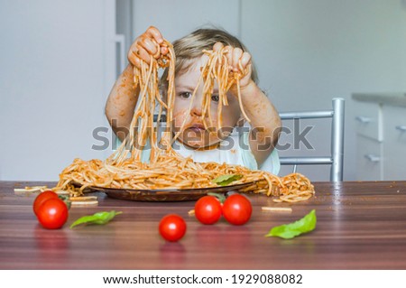 Funny baby child getting messy eating spaghetti with tomato sauce from a large plate, by itself with his hands, at home