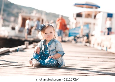 Funny Baby Girl 1-2 Year Old Sitting On Wooden Pier Wearing Denim Clothes Outdoors. Looking Away. Childhood. Summer Time. 