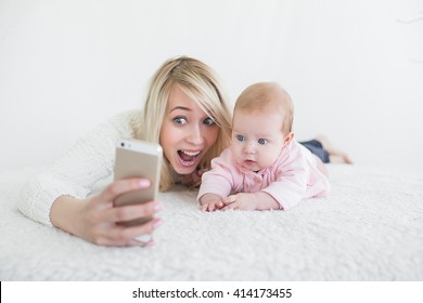 Funny Baby Girl Make Selfie On Mobile Phone And Lying Near Her Mother. Newborn Looking At The Camera And Smiling. 