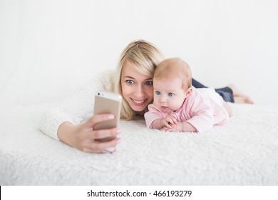 Funny Baby Girl Make Selfie On Mobile Phone And Lying Near Her Mother. Newborn Looking At The Camera And Smiling. 