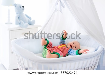Funny baby in white crib with canopy. Nursery interior and bedding for kids. Laughing little boy playing in moses basket. Bedroom with bassinet for young children. Happy child in colorful pajamas.