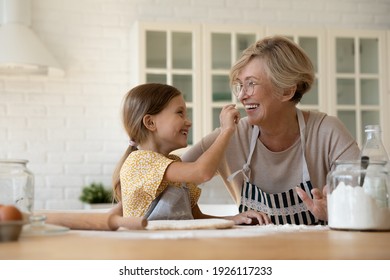 Funny Baking. Joyful Little Girl Having Fun With Retired Older Grandma When Cooking Pastries. Laughing Elderly Granny And Cute Small Grandchild Playing At Kitchen Painting One Another Noses With Flour