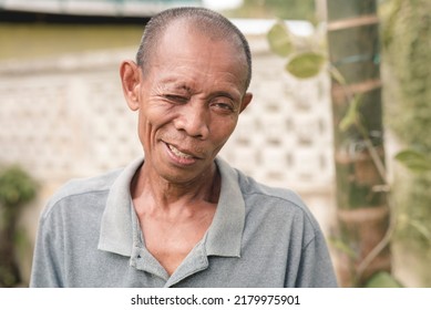 A Funny Bald Southeast Asian Man Makes An Awkward Wink Trying To Look Cute While Flirting Or Acknowledging An Inside Joke. Wearing A Gray Polo Shirt.