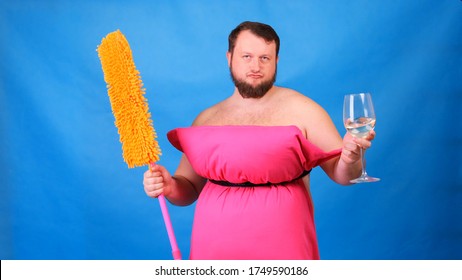 Funny Bearded Guy In A Pink Pillow Dress With A Mop And A Glass Of Wine On A Blue Background. Crazy Quarantine. Funny House Cleaning. Fashion 2020. Put On A Pillow. Challenge 2020 Due To House