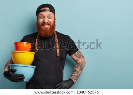 Funny bearded man clenches teeth, has cheerful expression, holds colourful utensils of different size, wears black headgear and apron, isolated in blue studio wall with empty space for information