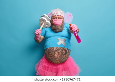 Funny Bearded Man With Fat Tattooed Belly Wears Fairy Costume Protective Mask Holds Detergent And Plunger Ready For Cleaning House Isolated Over Blue Background. Overweight Male Fairy Of Purity