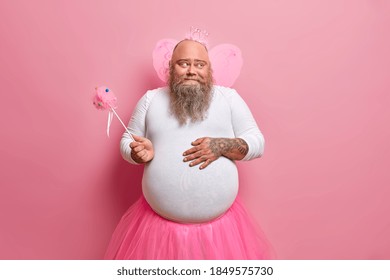 Funny bearded plump man keeps hand on fat belly holds magic wand looks happily aside entertains children at party isolated over pink background. Positive dad in carnival costume has playful mood