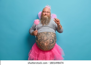 Funny Bearded Stout Man Dresses For Birthday Party, Wears Pink Crown, Pleated Skirt And Butterfly Wings, Takes Part In Magic Show Or Children Performance Isolated On Blue Background Makes Tiny Gesture