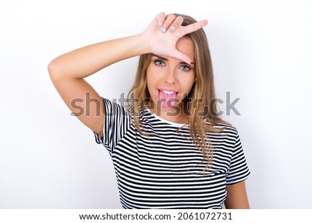 Funny beautiful blonde girl wearing striped t-shirt on white background makes loser gesture mocking at someone sticks out tongue making grimace face.