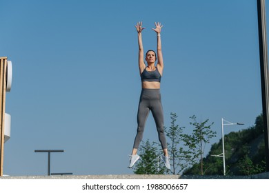 Funny Beautiful Brunette Exercising Outdoors, Jumping Vertically With Straight Body, Throwing Hands In The Air. In Grey Top And Yoga Pants. On A Sunny Day.