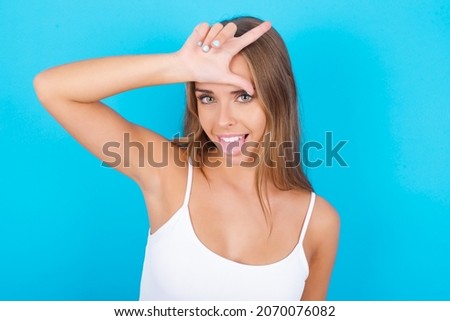 Funny Beautiful caucasian girl wearing white tank top over isolated background makes loser gesture mocking at someone sticks out tongue making grimace face.