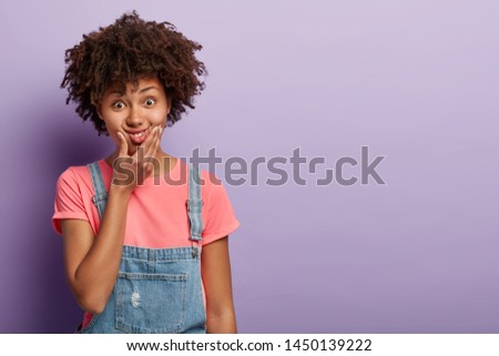 Funny beautiful woman with Afro hairstyle, presses lips, makes grimace, smiles and looks surprisingly, entertains child, tries be comic, wears casual outfit, poses over purple background, empty space
