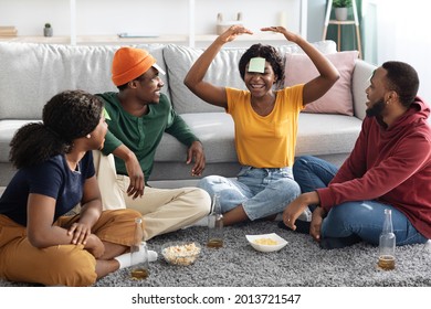 Funny black friends playing guess who game while relaxing together at home, sitting on floor in cozy living room, having cards on their foreheads, drinking beer, eating popcorn and laughing - Powered by Shutterstock