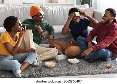 Funny black friends playing guess who game while chilling together at home, sitting on floor in cozy living room, having cards on their foreheads, drinking beer, eating popcorn and laughing