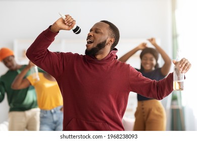 Funny Black Guy Singing Karaoke At Home, Holding Bottle Of Beer, Dancing And Cheering Group Of African American People On Background. Drunk Young Man Having Fun With His Friends