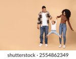 Funny black parents playing with daughter, holding hands and raising girl up, family posing over beige studio background, full length shot, copy space