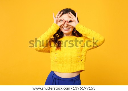 Funny blinking woman in sweater blue trousers hold hands near eyes imitating glasses or binoculars isolated on yellow orange background. People sincere emotions, lifestyle concept. Mock up copy space