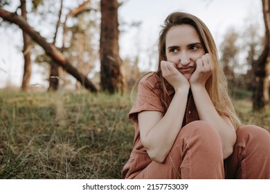 Funny Blonde Girl With Grumpy Face Putting Her Hands On Her Face And Sitting In Forest. Female Model With Comical And Ridiculous Face