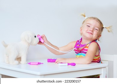 Funny blonde toddler girl playing doctor role game examinating her puppy using stethoscope sitting at small white table in playroom at home, school or kindergarten