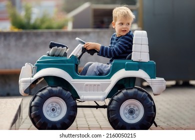 Funny Boy Car Driver With The Steering Wheel. Three Year-old Boy In A Blue Sweater In A Green Toy Car In The City. Little Boy Driving Big Toy Car And Having Fun, Outdoors.